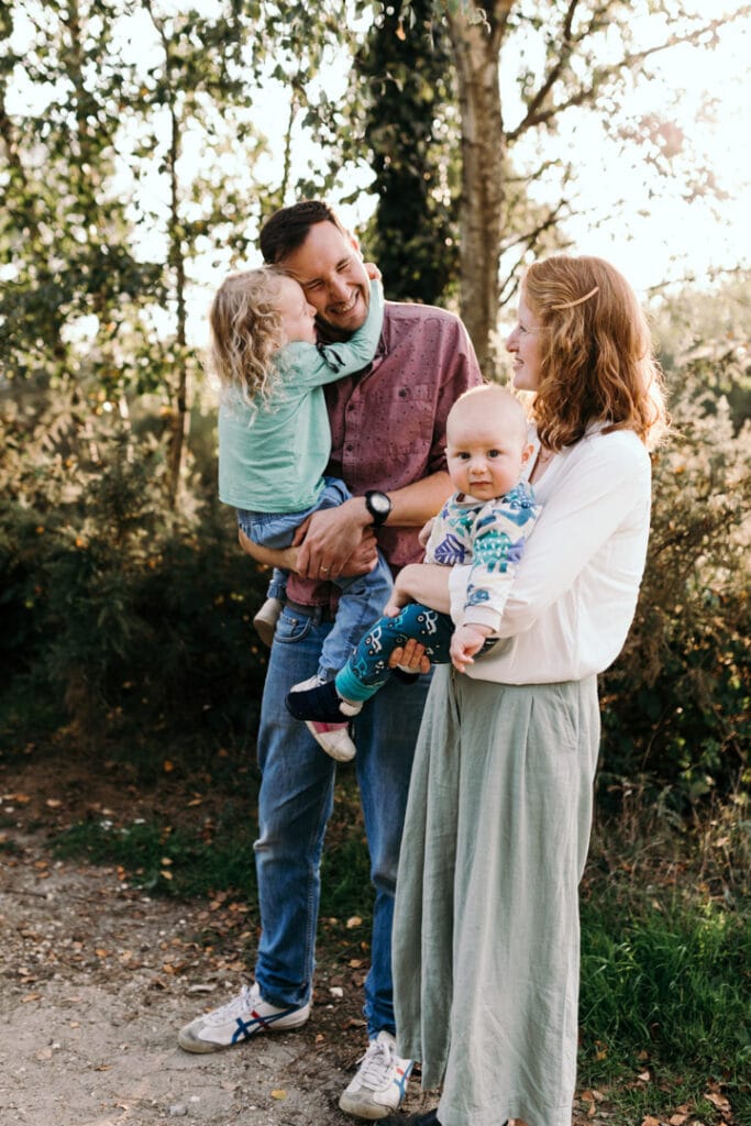 Family of four is standing and looking at each other. Older daughter is hugging her dad very close and mum is smiling and looking at her daughter. Little baby boy is looking at the camera. Family photographer in Hampshire. Ewa Jones Photography