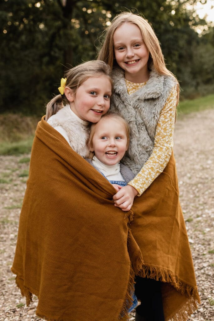 Three sisters are cuddling up inside the blanket. They are all smiling. Family photo session in Basingstoke, Hampshire. Ewa Jones Photography