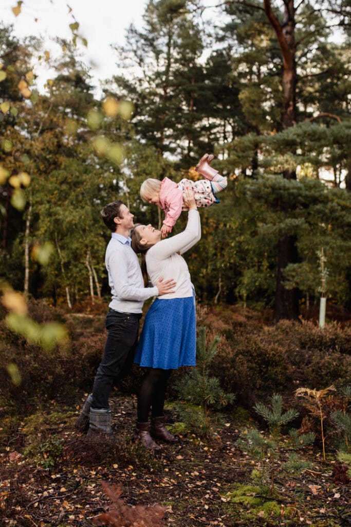 Mum is lifting into the air her little girl and dad is standing behind. Family autumnal photo session in Surrey. Family photographer. Ewa Jones Photography