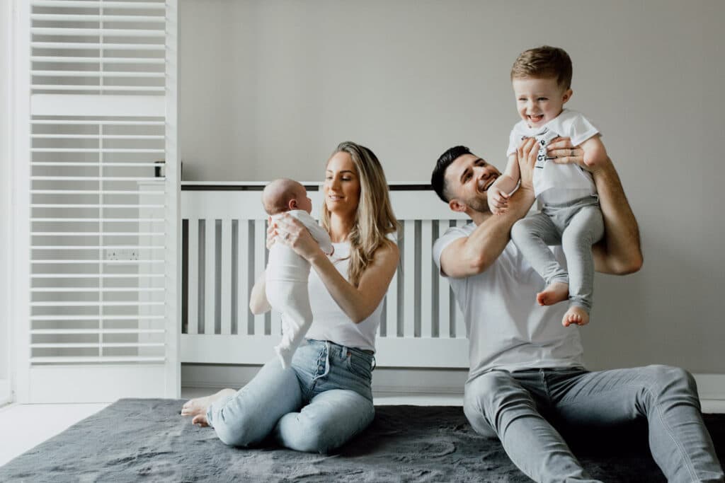 Mum and dad are sitting on the floor next to the window. They are both holding their children up and smiling. They are all wearing white tops and blue jeans. Family photographer in Basingstoke, Hampshire. Ewa Jones Photography