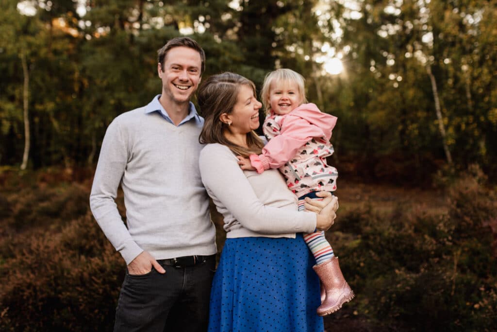 Pregnant mum is holding her toddler girl and looking at her. Dad and little girl are looking at the camera. Lovely family photo session in Surrey. Ewa Jones Photography