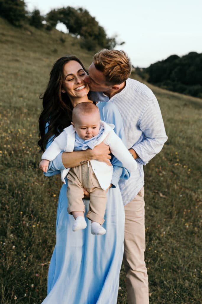 Dad is hugging mum and kissing her on her cheek. Mum is holding her six month old baby. They are all wearing light blue clothing. Family photographer in Hampshire. Ewa Jones Photography