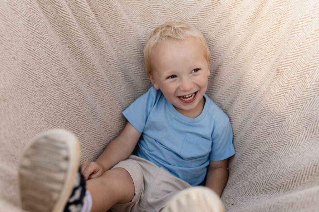 Little boy is inside the blanket and smiling. He is wearing blue top and cream shorts. Candid family photography in Hook, Hampshire. Ewa Jones Photography