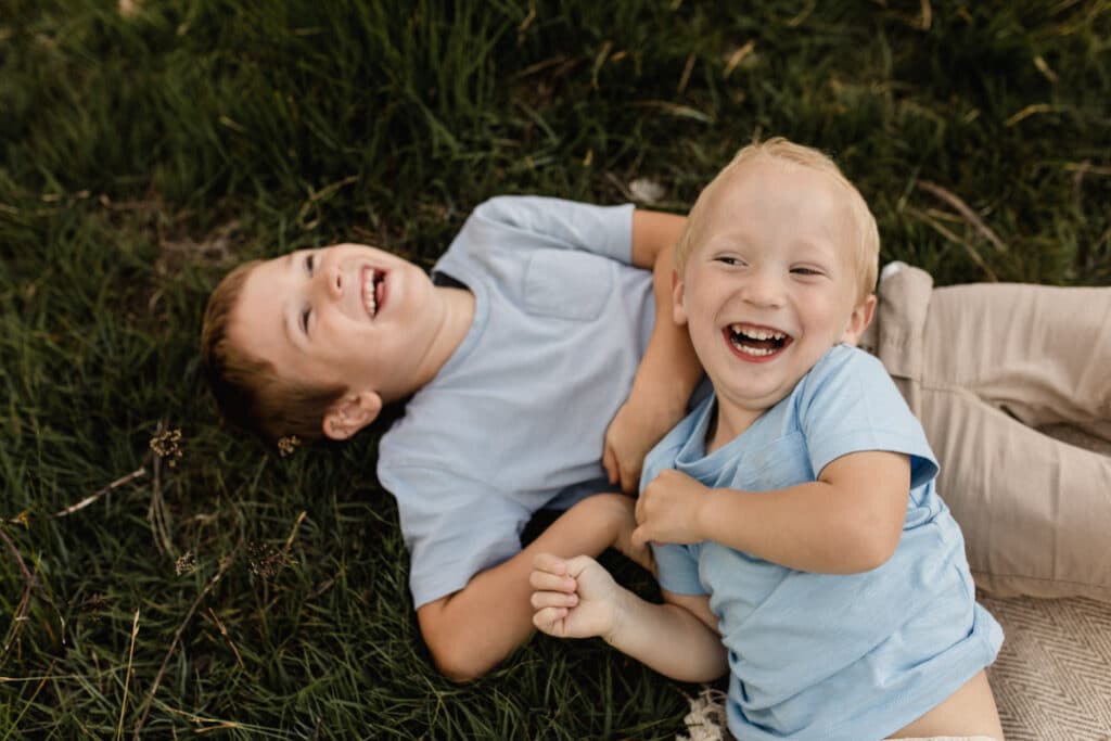 One brother is laying on the grass laughing and another brother is laying on his sibling. Younger brother is laughing too. Family photographer in Hampshire. Ewa Jones Photography