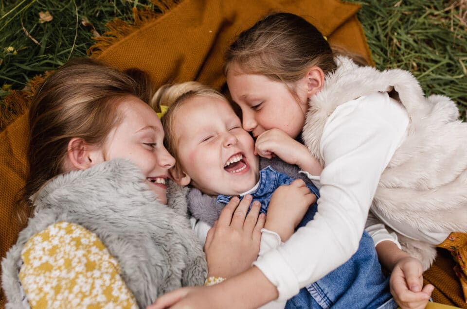 Three sisters are laying on the blanket and hugging each other. They are all wearing lovely autumnal clothing. Golden hour autumn photo session in Basingstoke, Hampshire. Ewa Jones Photography