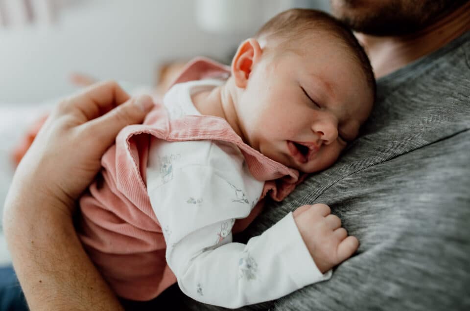 Newborn baby girl is sleeping on daddy. She is wearing lovely white baby grow and pink dress. Newborn baby photographer in Hampshire. Ewa Jones Photography