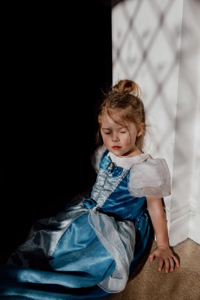Little girl is sitting on the floor and has her back against the wall. She has her eyes closed and dreaming. She is wearing a Cinderella dress. Family photographer in Hampshire. Ewa Jones Photography