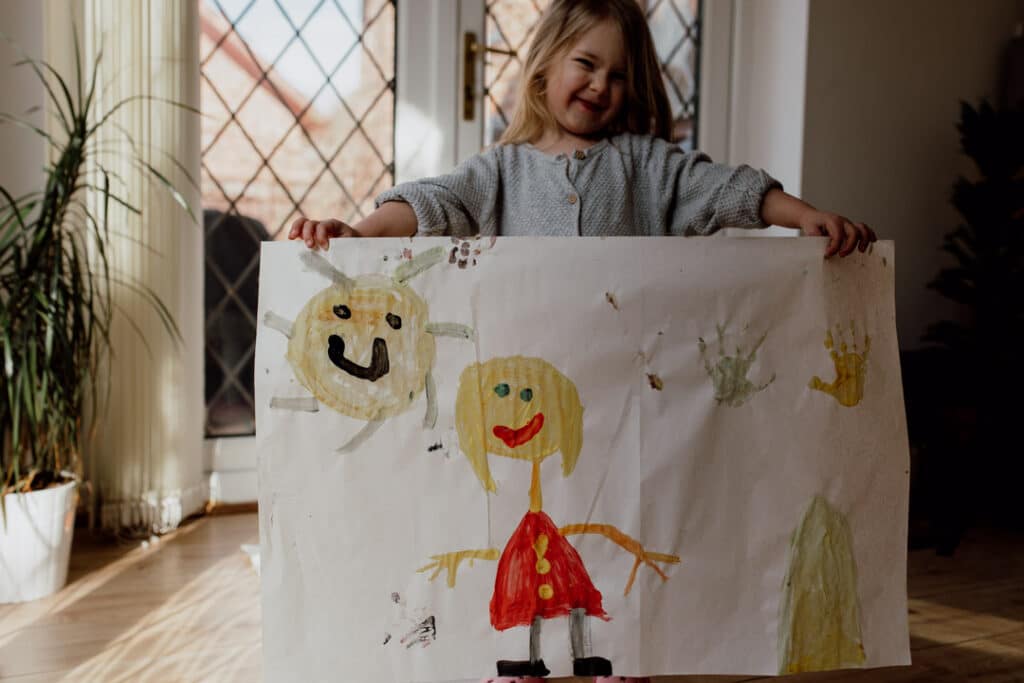 Little girl is holding a big picture that she has painted. It is a picture of a girl, the sun, house and hand prints. Family lifestyle photographer in Hampshire. Ewa Jones Photography