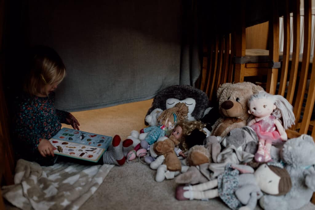 Little gir is sitting under a den and reading a book to all her cuddly toys. Natural family lifestyle photography in Hampshire. Ewa Jones Photography