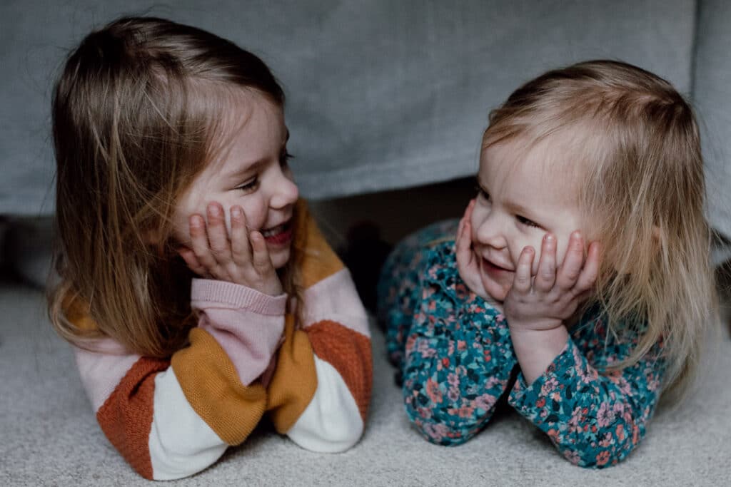 Two sisters are laying on the floor and looking at each other. They are both smiling to each other. 7 tips on photographing kids at home. Family photography in Hampshire. Ewa Jones Photography