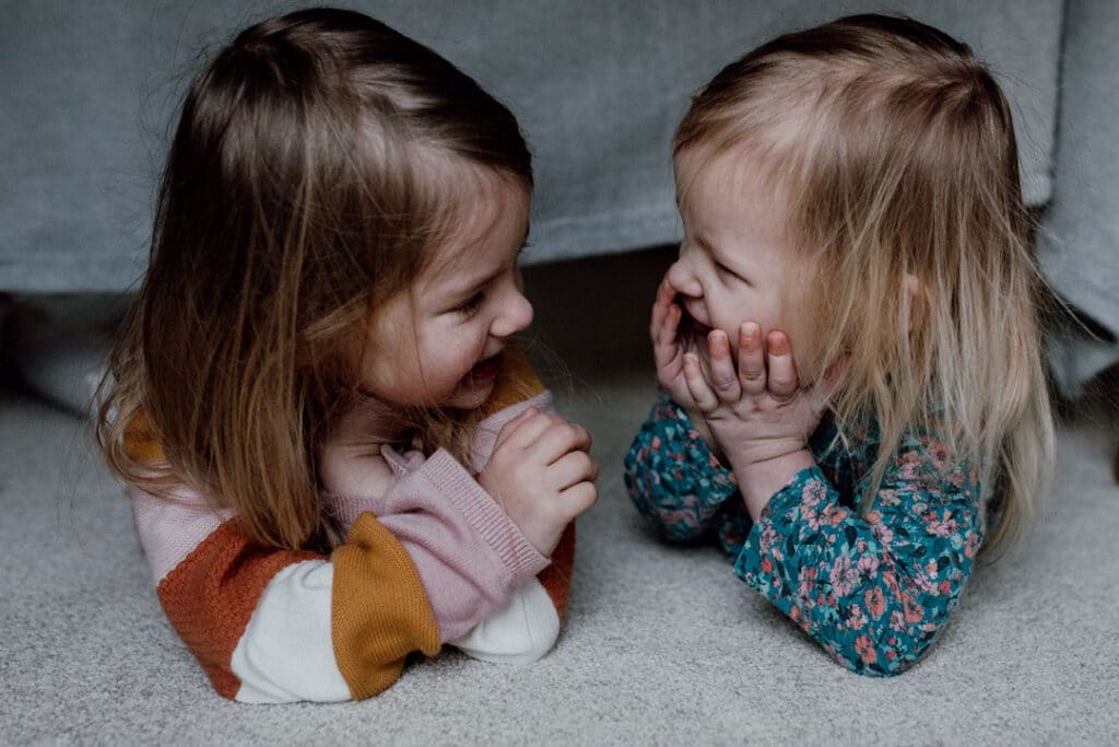 Two sisters are laying on the floor and looking at each other. They are both smiling to each other. 7 tips on photographing kids at home. Family photography in Hampshire. Ewa Jones Photography