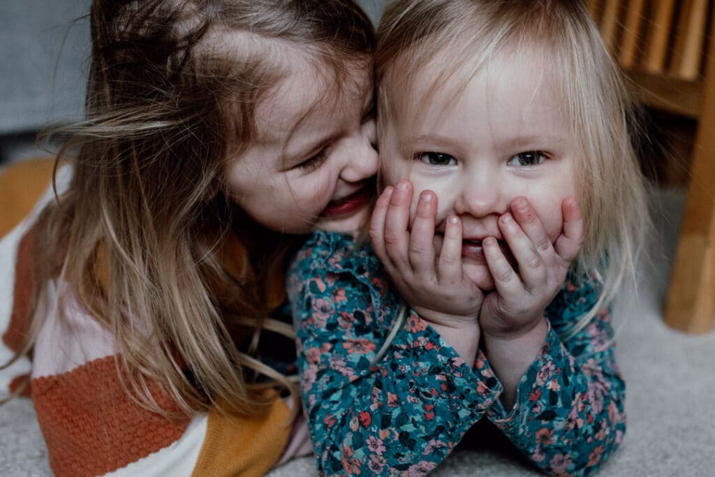 Two sisters are laying on the floor. Younger sister has her hands on her face and looking at the camera. Other sister is looking at her and cuddling her. Family photographer in Hampshire. Ewa Jones Photography