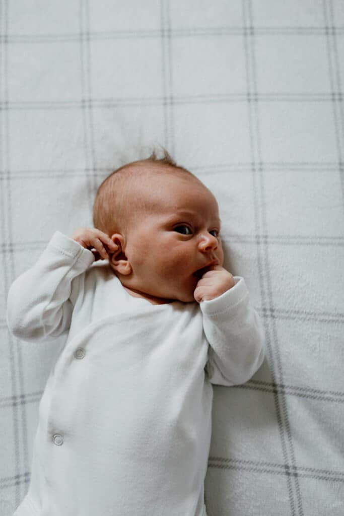 Newborn baby boy is laying on the bed and he is wearing a white baby grow. Newborn baby photo shoot in Hampshire. Ewa Jones Photography