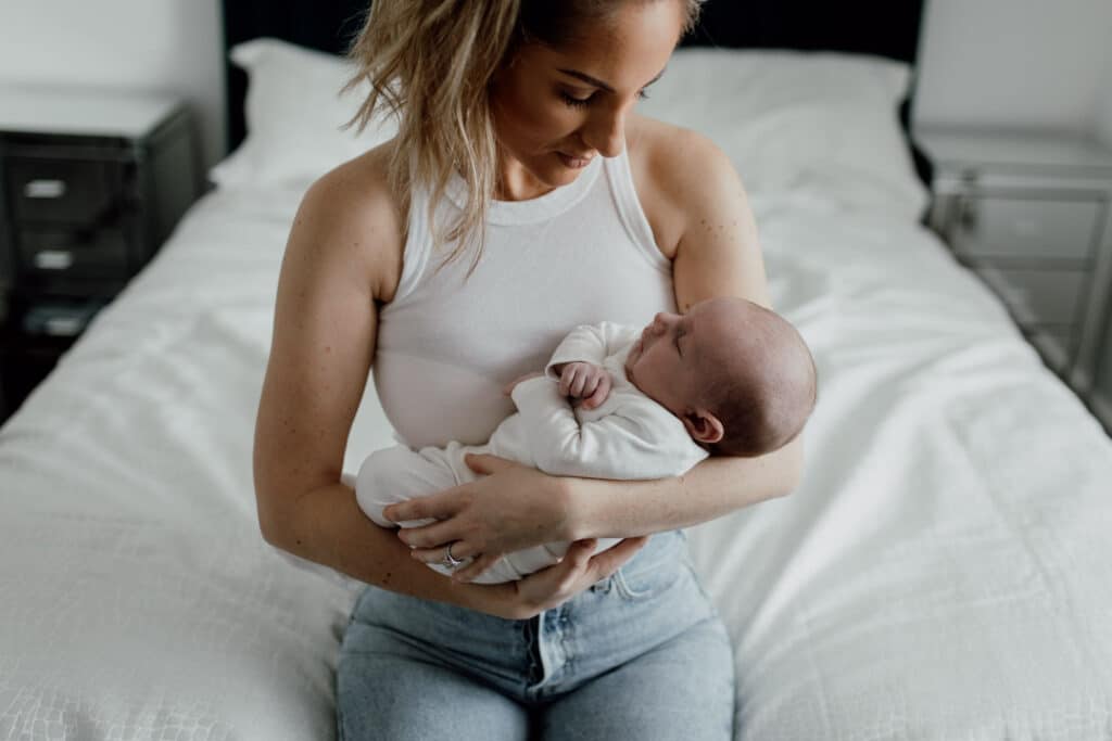 mum is holding her newborn baby. Newborn baby is sleeping and he is wearing a white baby grow. Newborn baby photographer in Hampshire. Ewa Jones Photography