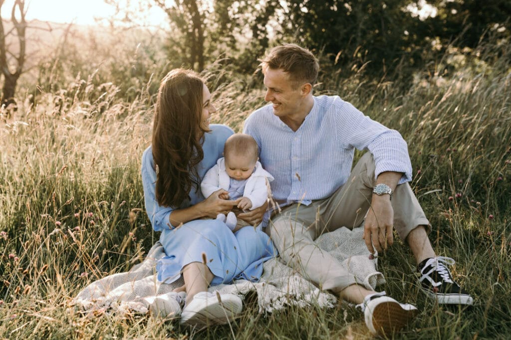 Family of three is sitting on the grass. mum and dad are looking at each other and smiling. Family photo session in Hampshire. Ewa Jones Photography