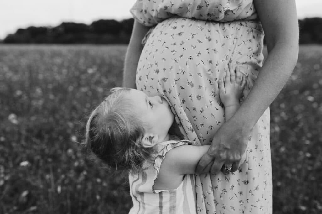 Little girl is kissing her mum bump and looking up at her mum. Black and white family photography. Family photographer in Basingstoke, Hampshire. Ewa Jones Photography