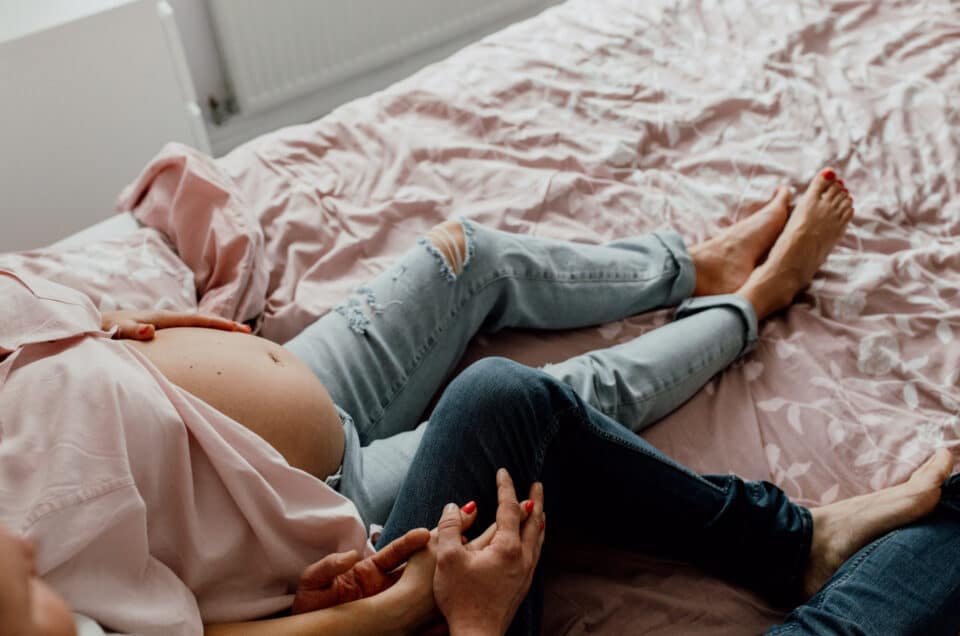 Expecting mum is sitting on the bed and she holds her hands with her husband. Lovely candid in home maternity photo shoot in Hampshire. Ewa Jones Photography