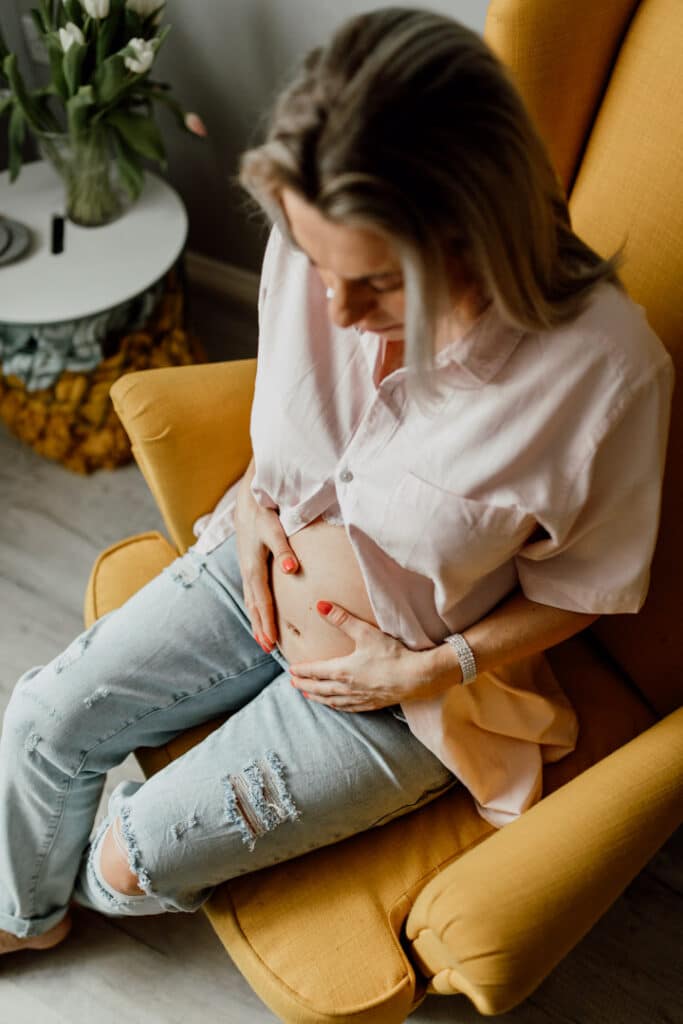 Pregnant mum is sitting on the yellow chair and looking down at her bump. Maternity photoshoot in Hampshire. Ewa Jones Photography