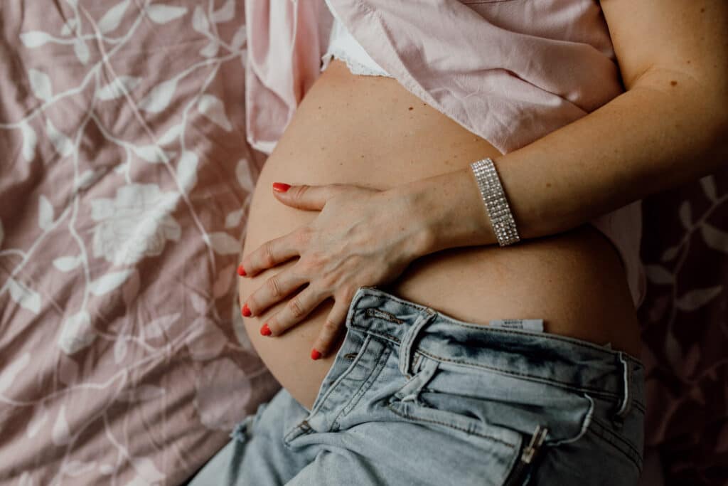 Pregnant mum is laying on the bed and holding her baby bump. She is wearing a pink shirt and jeans. Maternity photo session in Hampshire. Ewa Jones Photography