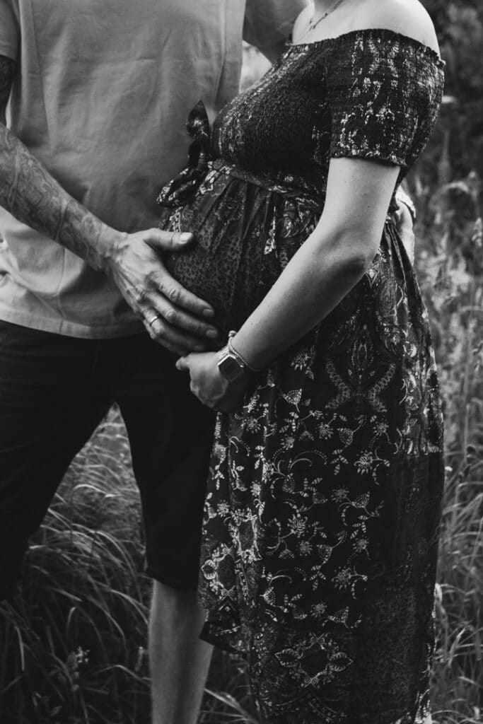 Expecting mum is standing with her hand on her baby bump. Dad is also holding baby bump. Black and white maternity photograph. Maternity photographer in Hampshire. Ewa Jones Photography