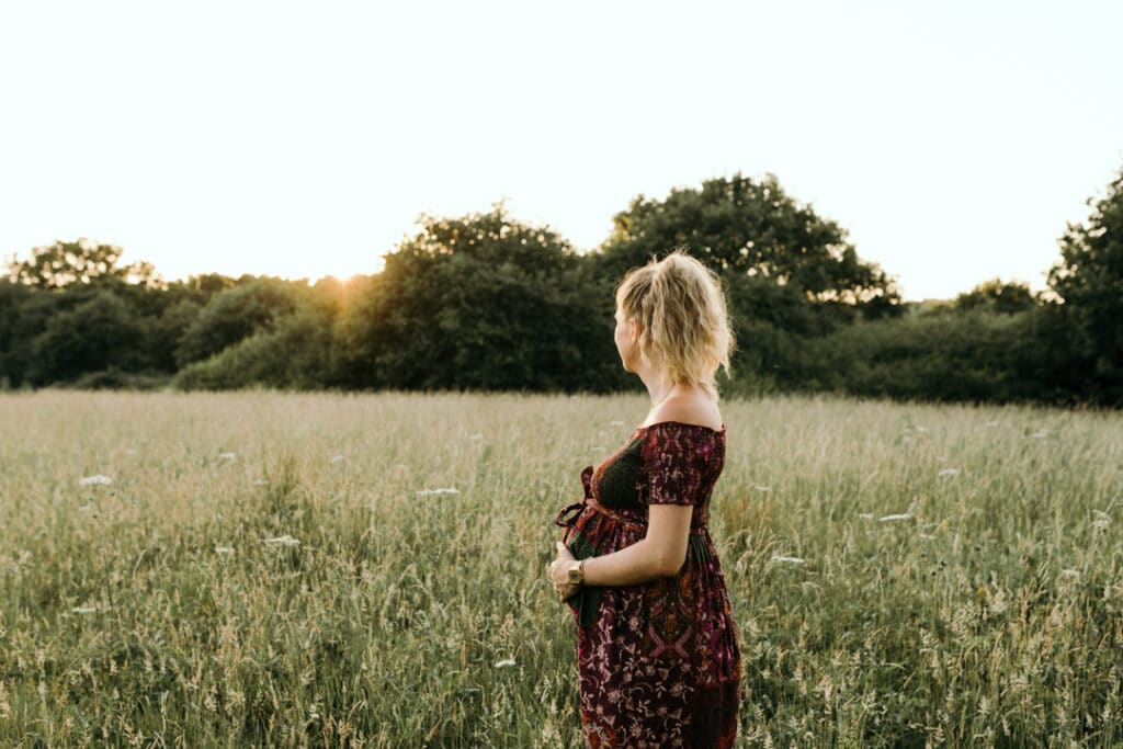 Pregnant mum is looking at the fields. She is holding her baby bump and wearing lovely flower dress. Maternity photographer in Hampshire. Ewa Jones Photography
