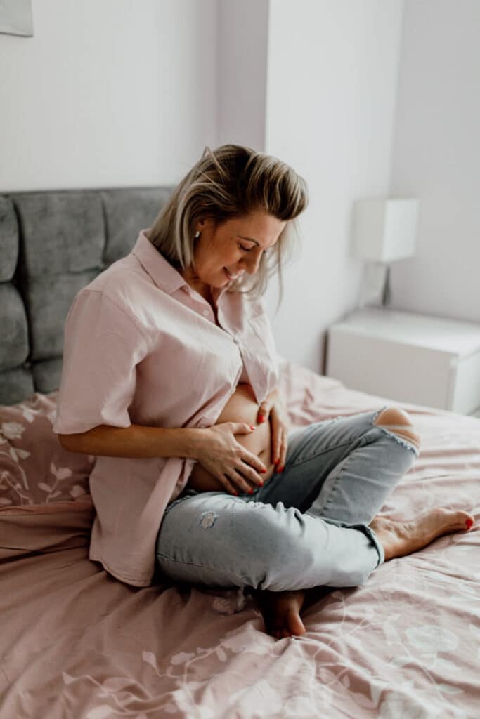 Expecting mum is sitting cross leg on the bed and holding baby bump. she is looking down at the bump. She is wearing jeans and a pink shirt. Maternity photographer in Hampshire. Ewa Jones Photography