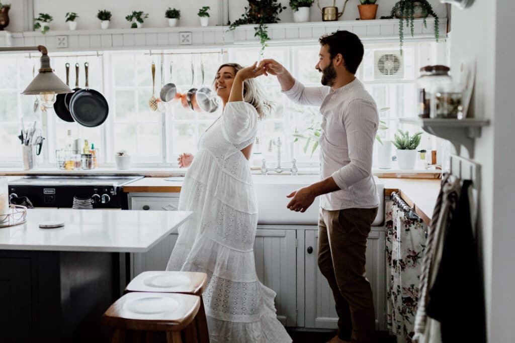 Expecting parents are dancing in the kitchen. Mum is wearing a lovely maternity dress. in home maternity photo session in Hampshire. Ewa Jones Photogrraphy