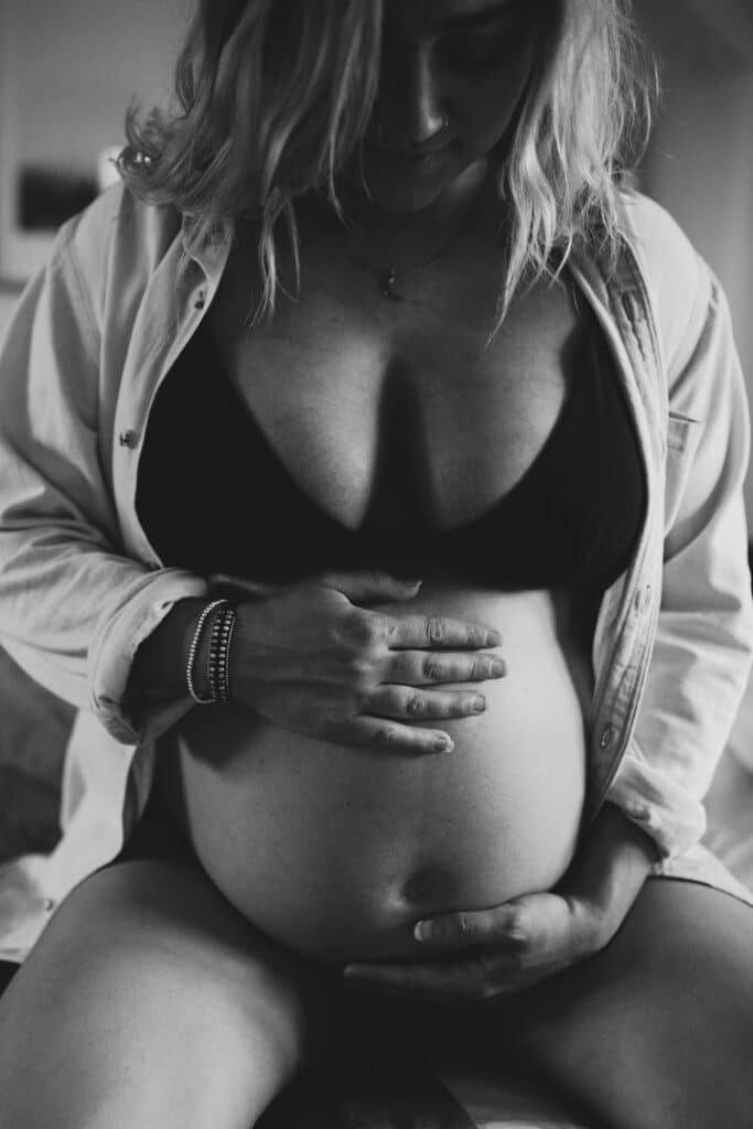 Black and white photography of mum sitting on the bed and holding her bump. Lovely maternity photography at home. Family photographer in Hampshire. Ewa Jones Photography