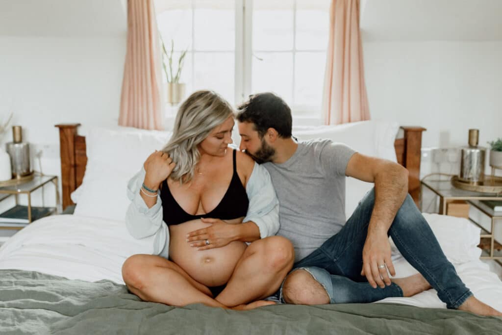 Mum and dad are sitting on the bed. Mum is holding her bump and dad is kissing expecting mum on her shoulder. Maternity photographer in Hampshire. Ewa Jones Photography