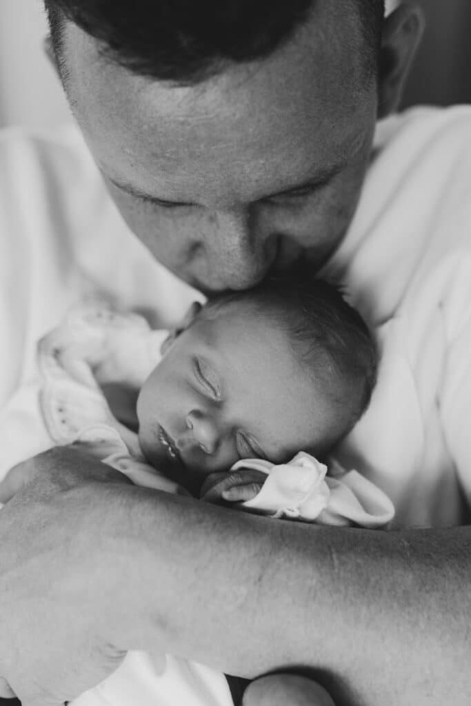 dad is kissing his newborn baby girl and holding her close to his face. Newborn baby girl is sleeping. Newborn photographer in Aldershot. Ewa Jones Photography