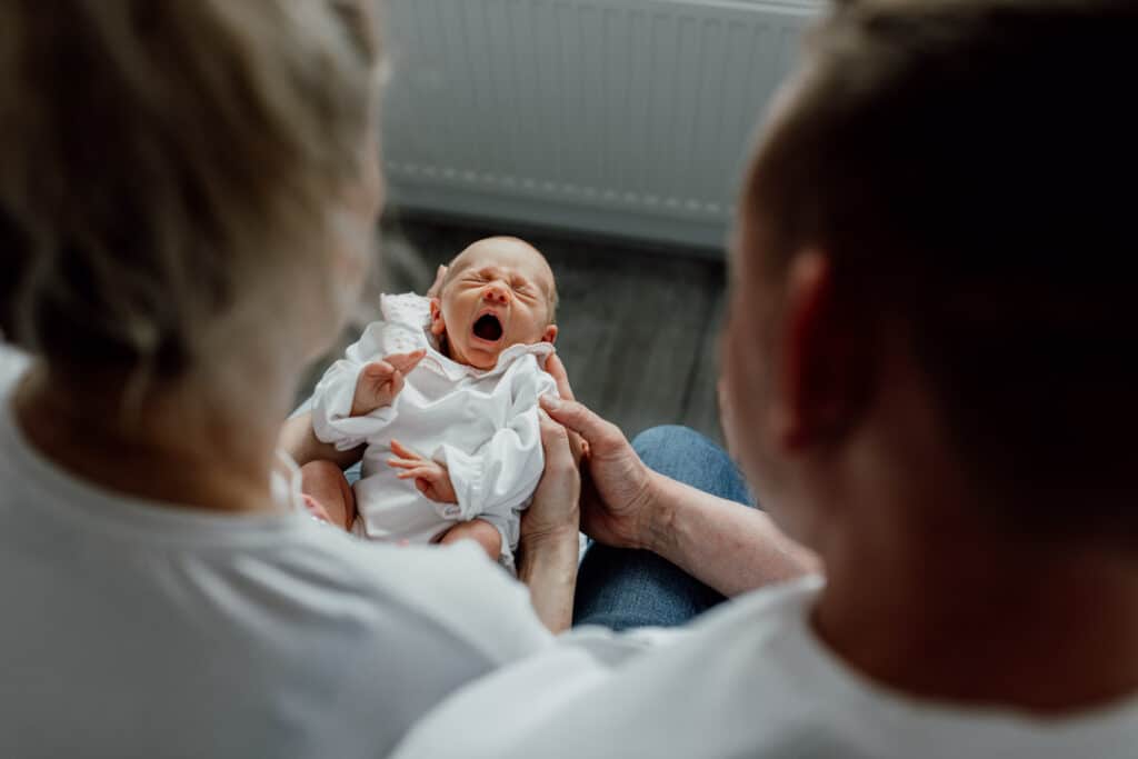 Mum and dad are looking down at their newborn baby girl who is yawning. Lovely natural in-home lifestyle newborn photo session. Ewa Jones Photography
