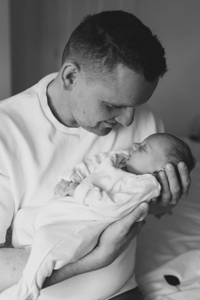 Black and white photograph of dad holding his newborn baby girl. Newborn baby girl is sleeping and dad is holding her close to him. Newborn baby photographer in Aldershot, Hampshire. Ewa Jones Photography