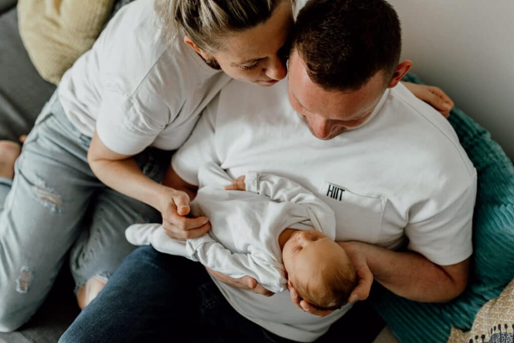 Mum and dad are sitting on the sofa and looking down at their newborn girl. Dad is holding his baby and she is sleeping. Lovely natural lifestyle newborn photography in Hampshire. Ewa Jones Photography