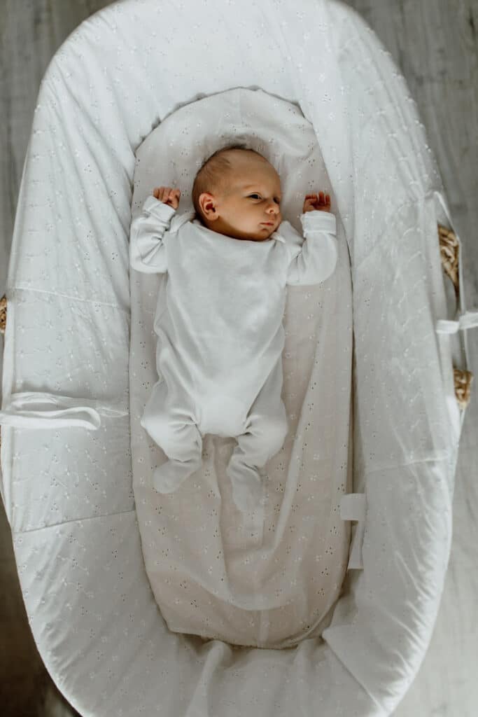 Newborn baby girl is laying in her moses basket. She is wearing a white body and has her hands up. Natural in-home newborn baby photography in Hampshire. Ewa Jones Photography