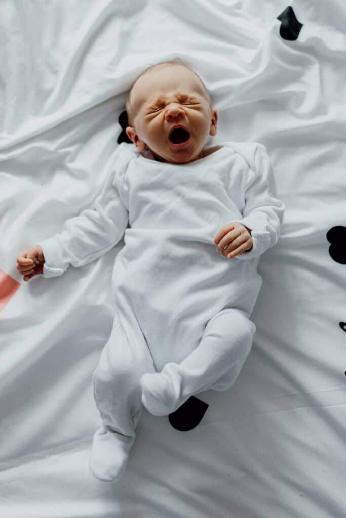 Newborn baby is wearing white baby grow and she is laying on the bed yawning. in-home lifestyle newborn baby photo session in Hampshire. Ewa Jones Photography