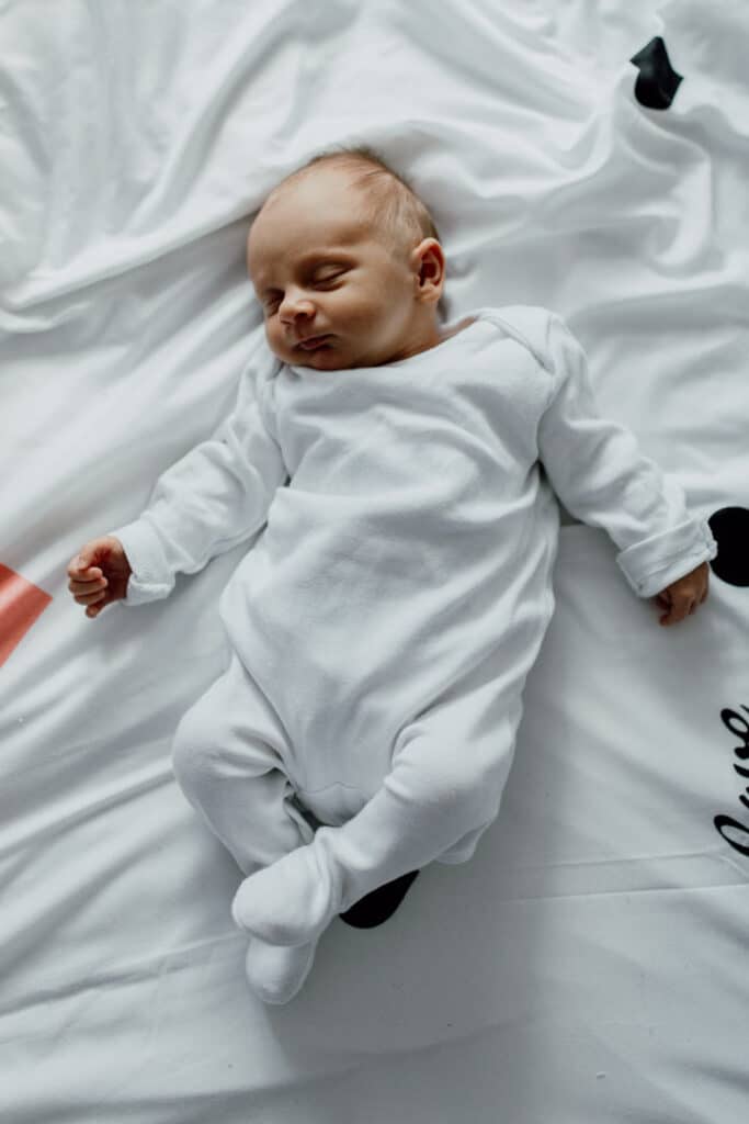 Newborn baby is wearing white baby grow and she is laying on the bed sleeping. in-home lifestyle newborn baby photo session in Hampshire. Ewa Jones Photography