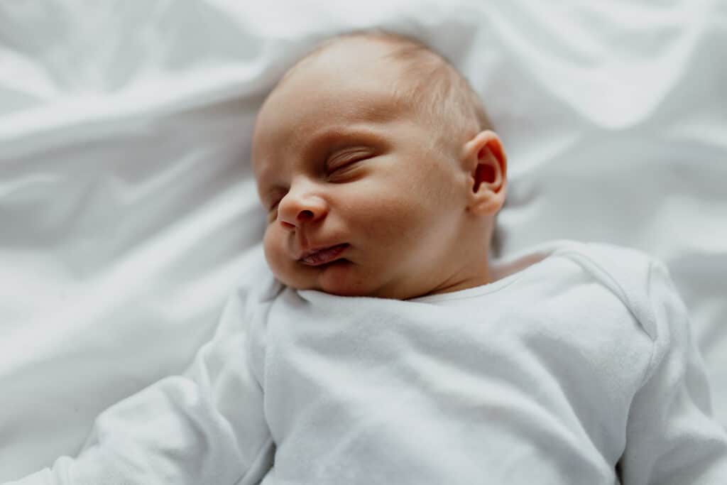 Newborn baby is wearing white baby grow and she is laying on the bed sleeping. in-home lifestyle newborn baby photo session in Hampshire. Ewa Jones Photography