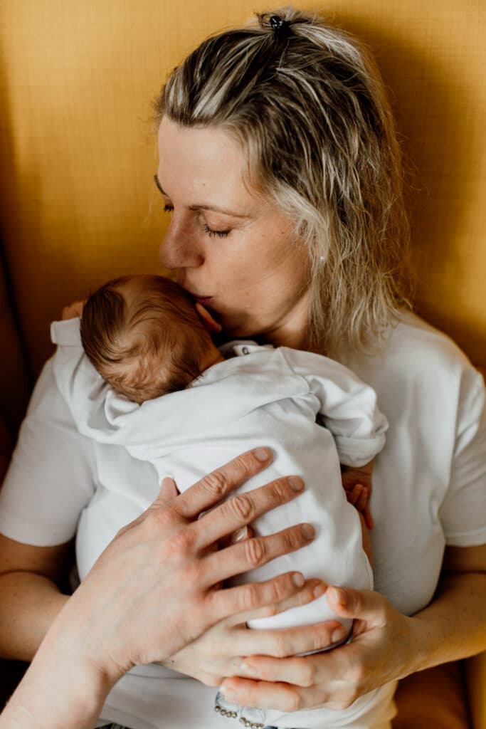 Mum is sitting on the yellow chair and holding her newborn baby girl. She is kissing the baby on her head. Dad is also holding his hand on baby's back. Newborn photographer in Hampshire. Ewa Jones Photography