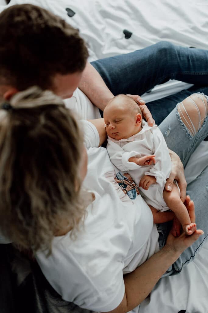 Mum and dad are both holding a newborn baby. They are sitting on the bed and looking down at the newborn baby. Newborn photographer in Basingstoke. Ewa Jones Photography