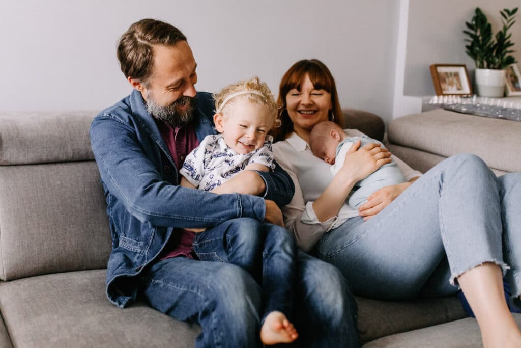 Lifestyle photograph of family sitting on the sofa and mum is cuddling her newborn baby boy and dad is hugging their older son.Newborn photoshoot in Hampshire. Ewa Jones Photography