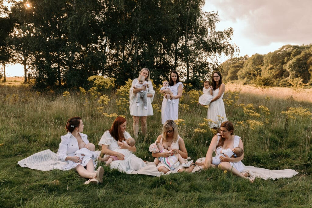 Mums are sitting on the blanket and breastfeeding their babies. There is a gorgeous field full of wild flowers and the sun is shining through the trees. Group breastfeeding photo session in Basingstoke, Hampshire. Ewa Jones Photography