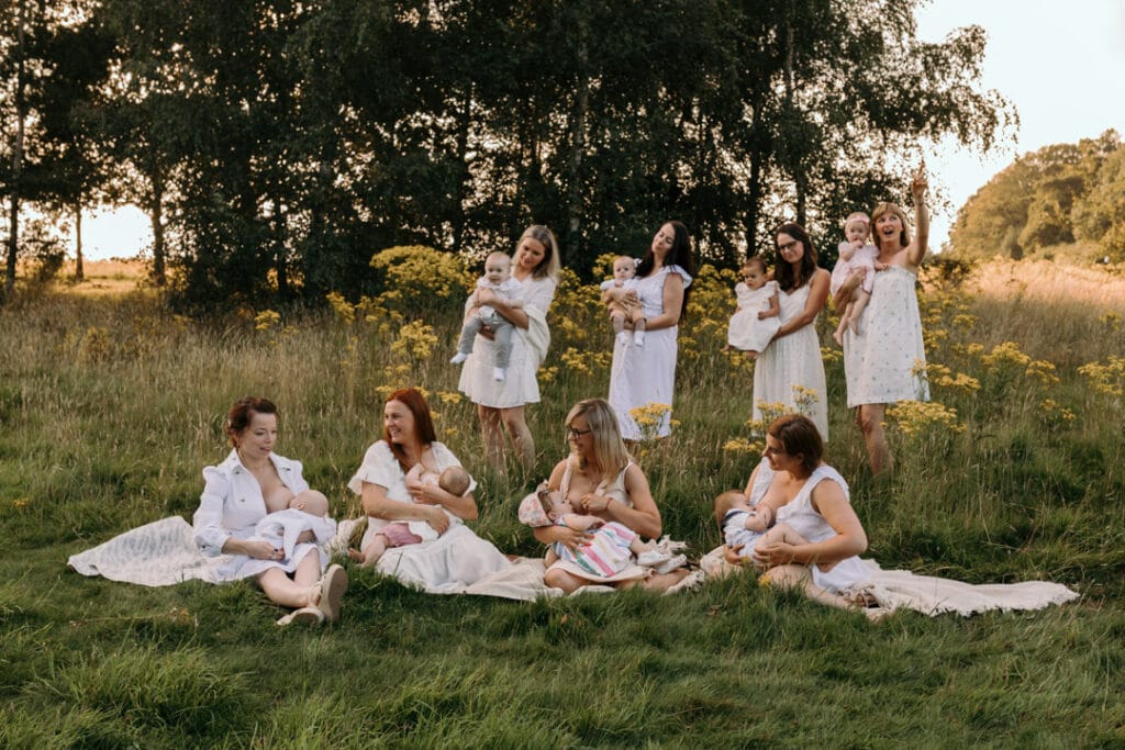 mums are sitting on the blanket and breastfeeding their babies. There is a gorgeous field full of wild flowers and the sun is shining through the trees. Group breastfeeding photo session Basingstoke, Hampshire. Ewa Jones Photography