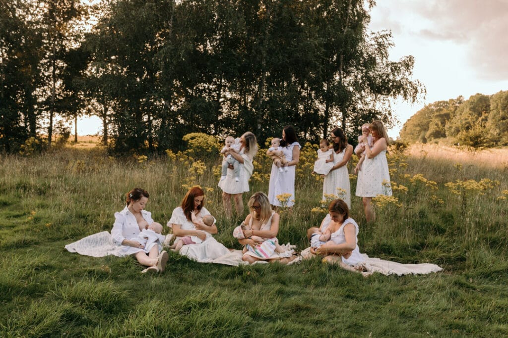 mums are sitting on the blanket and breastfeeding their babies. There is a gorgeous field full of wild flowers and the sun is shining through the trees. Group breastfeeding photo session Basingstoke, Hampshire. Ewa Jones Photography