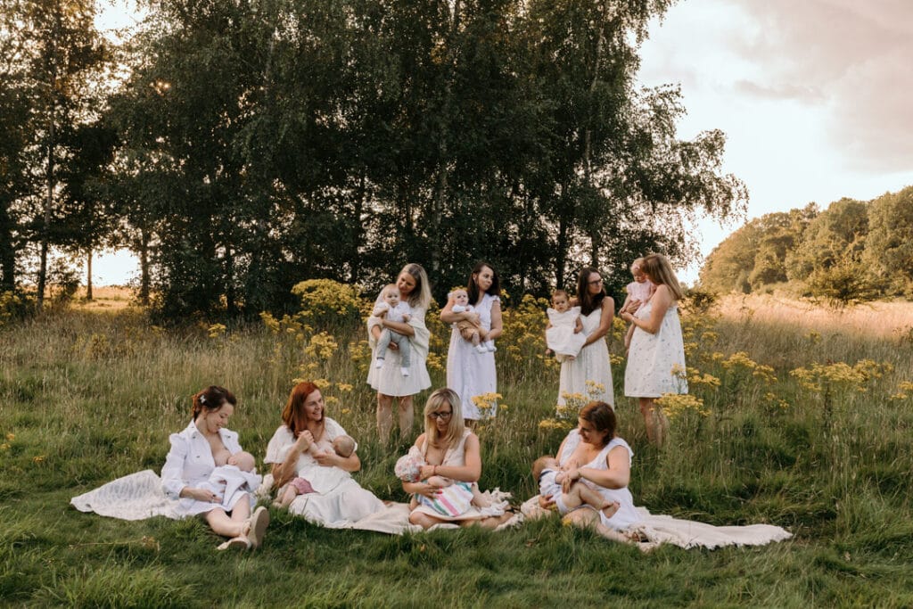 mums are sitting on the blanket and breastfeeding their babies. There is a gorgeous field full of wild flowers and the sun is shining through the trees. Group breastfeeding photo session Basingstoke, Hampshire. Ewa Jones Photography