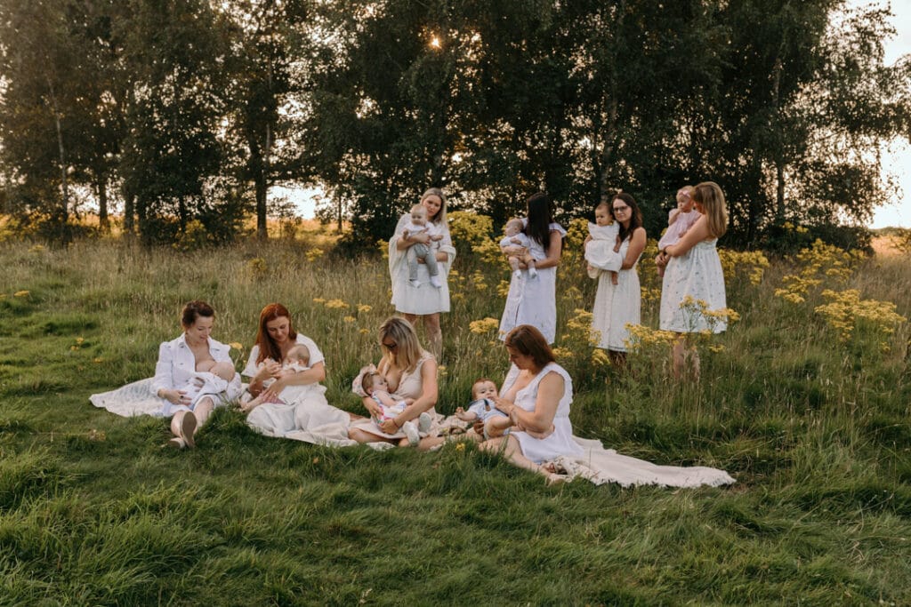 mums are sitting on the blanket and breastfeeding their babies. There is a gorgeous field full of wild flowers and the sun is shining through the trees. Group breastfeeding photo session in Basingstoke, Hampshire. Ewa Jones Photography