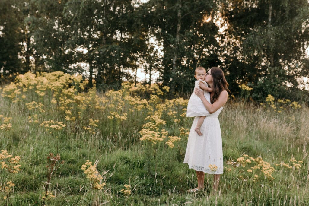 Mum is standing on the field of wild flowers and holding her girl. Mum is kissing her little girl on the cheek. Lovely family photography in Hampshire. Ewa Jones Photography