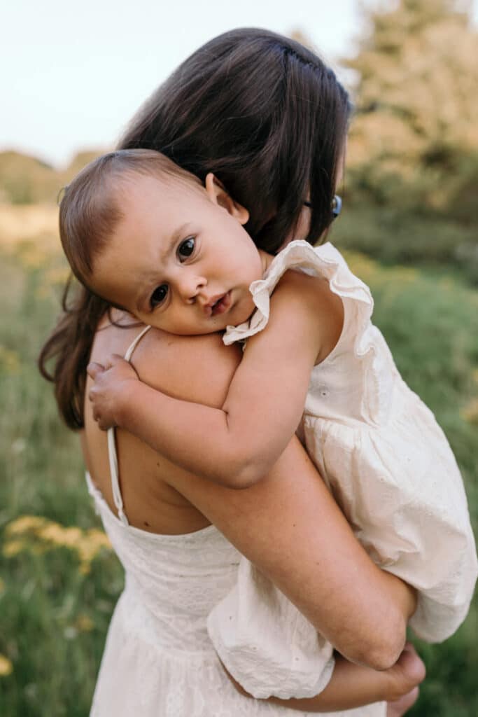 Little girl is laying gently on mummys arms. She is looking at the camera. Little girl is wearing lovely white dress. Family photographer in Basingstoke, Ewa Jones Photography