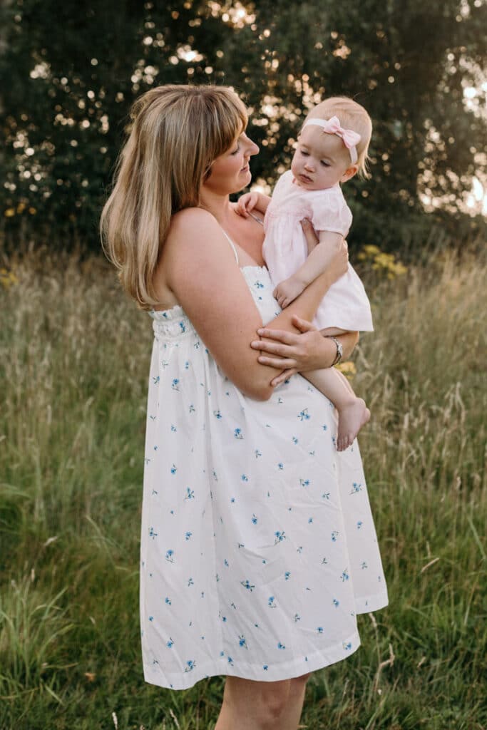 Mum is holding her little girl in her arms a looking at her. Sun is shining and mum is wearing white dress with blue flowers and girl is wearing lovely pink dress. Family photographer in Basingstoke, Hampshire. Ewa Jones Photography