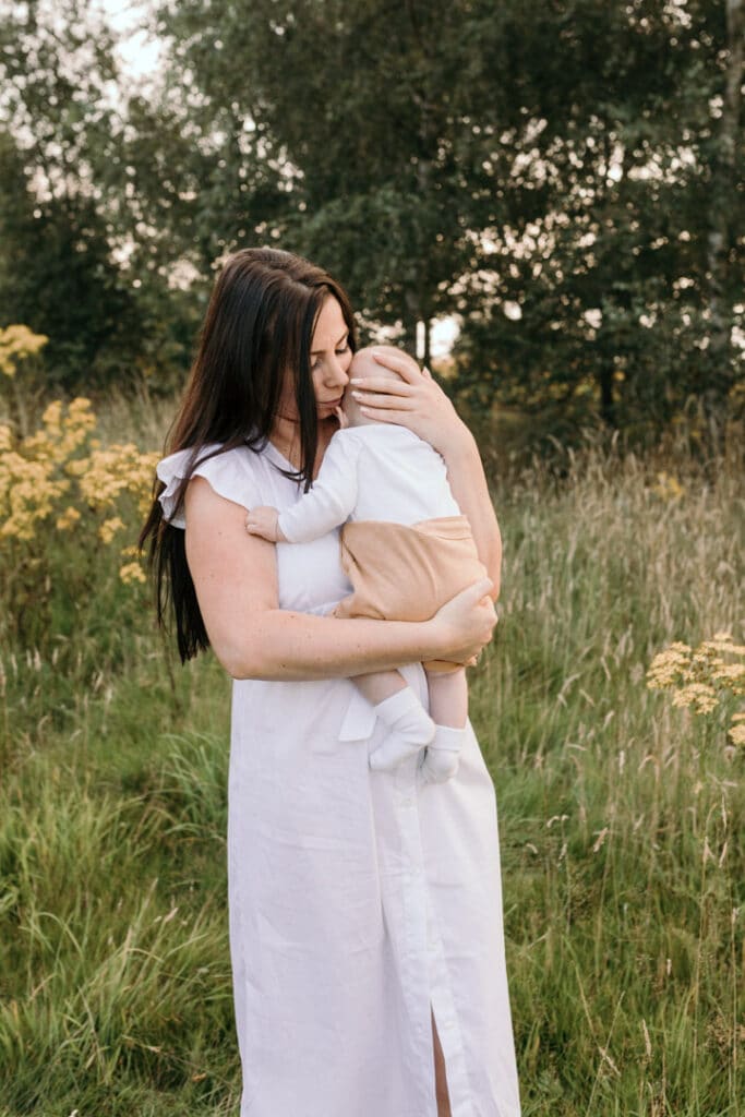 Mum is holding close her little baby boy and standing in the field of flowers. Sunset photo session. Mum is lifting up her little baby boy and he is looking at the camera and smiling. Sunset photo session in Basingstoke. Family photographer in Hampshire. Ewa Jones Photography