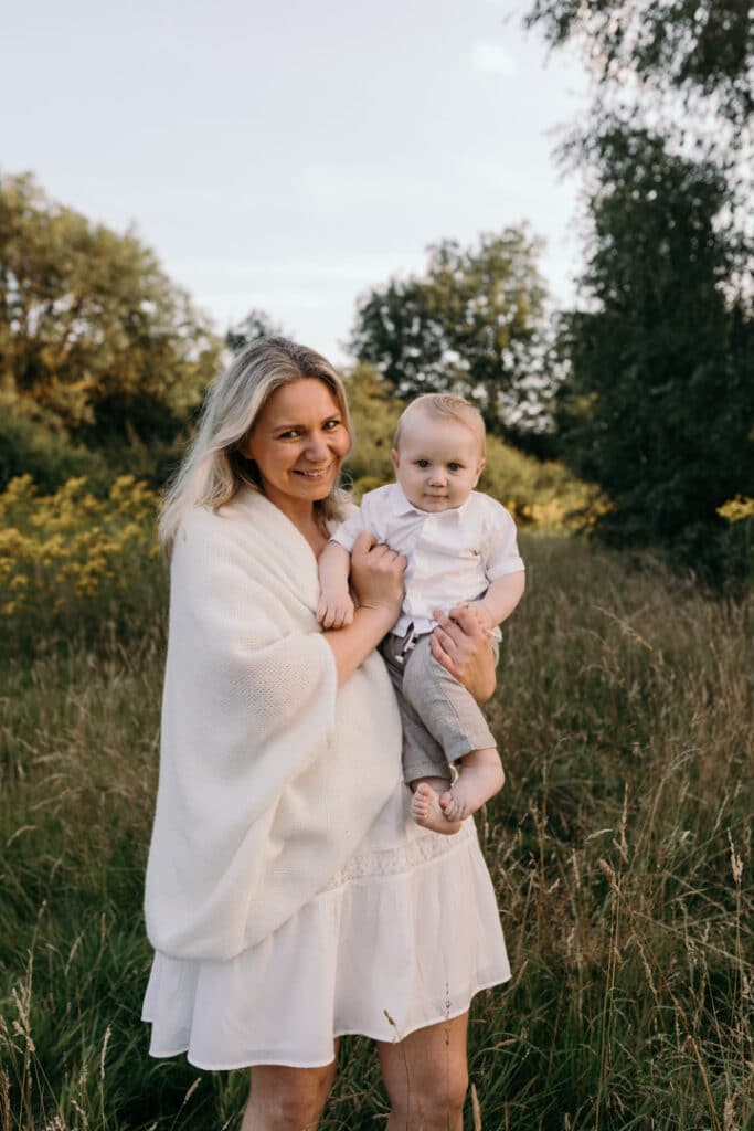 Mum is holding her baby boy and smiling. Mum is wearing lovely white dress and boy is wearing white shirt and grey trousers. Family photography in Basingstoke, Hampshire. Ewa Jones Photography